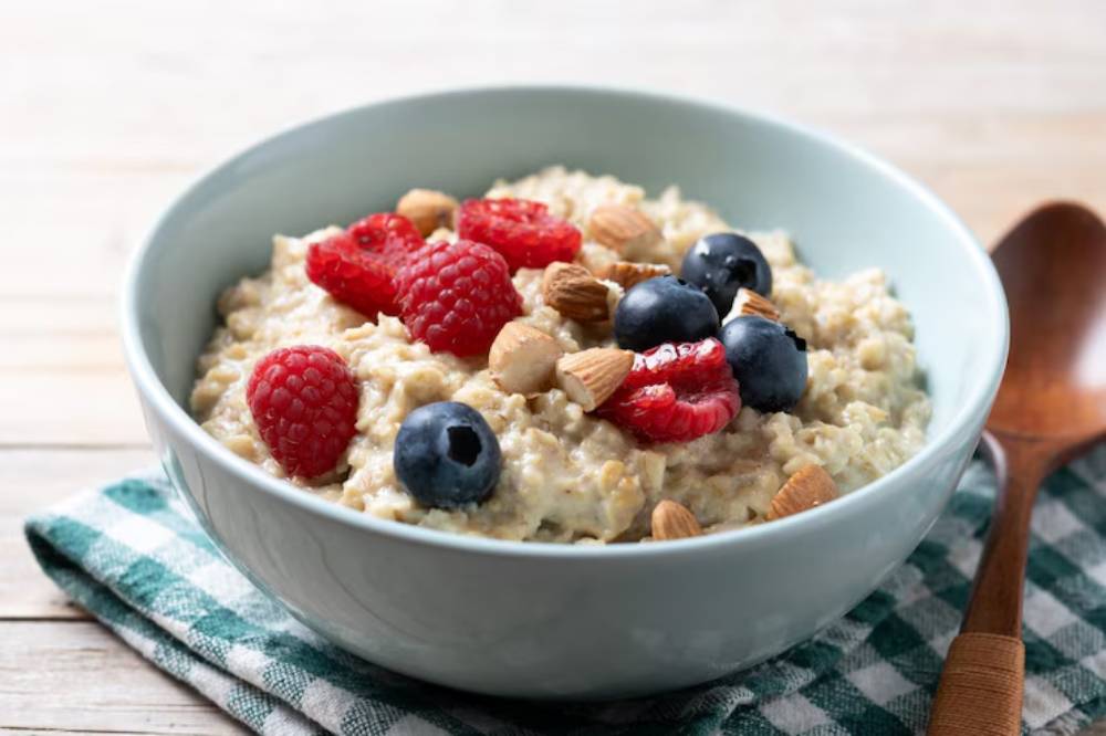 A bowl of oatmeal topped with raspberries, blueberries, and almonds, resting on a green checkered cloth next to a wooden spoon.