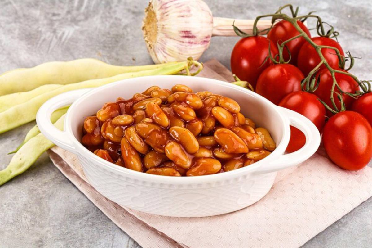 A bowl of baked beans in sauce, surrounded by fresh garlic, tomatoes, and green beans on a textured surface.