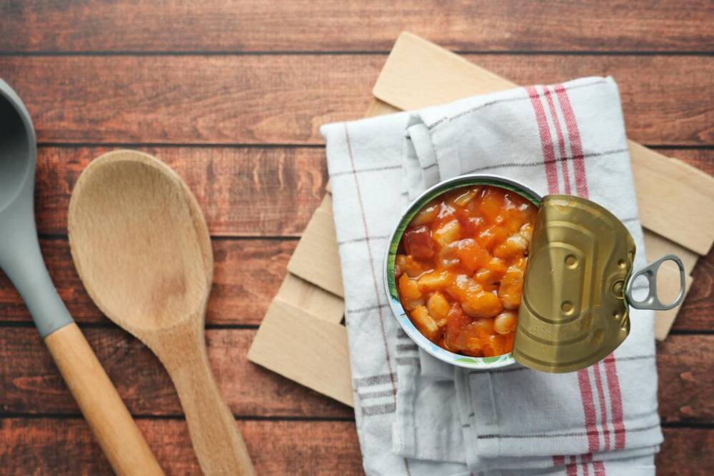 Open can of mixed beans in tomato sauce on a checkered cloth, surrounded by wooden utensils on a rustic wooden surface.