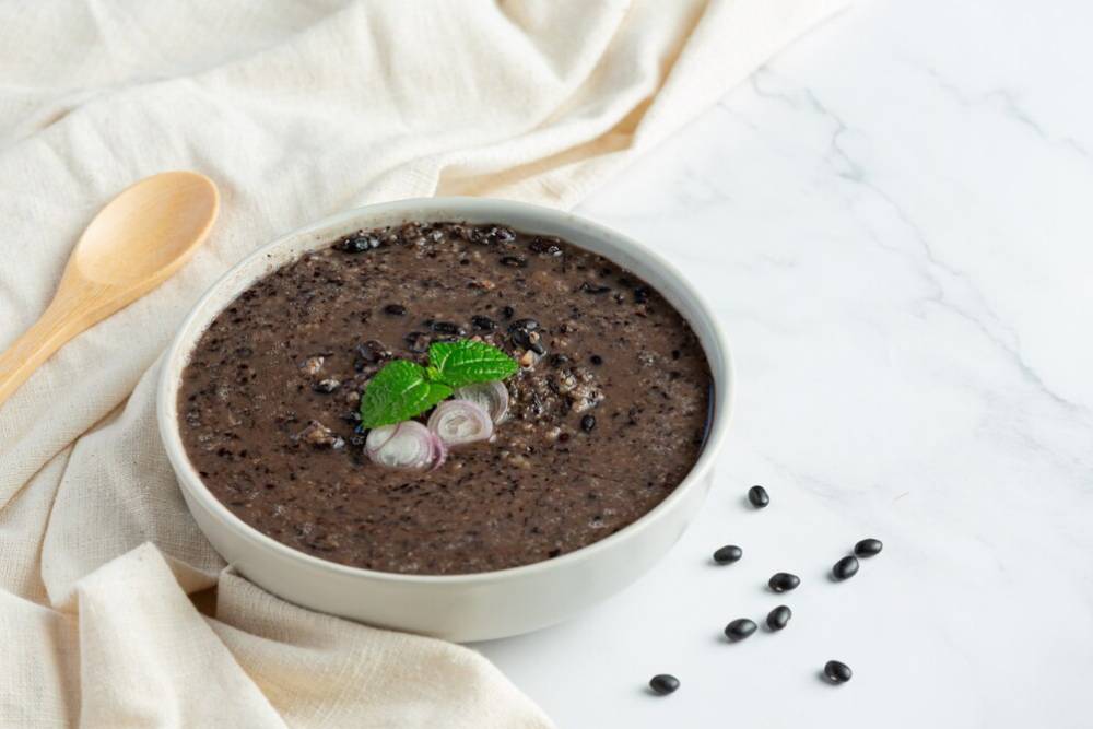 Bowl of black bean soup garnished with mint and sliced onions, surrounded by black beans and a wooden spoon on a soft linen backdrop.