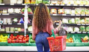 A woman with curly hair stands in a grocery store, holding a shopping basket filled with vegetables and flowers, browsing the produce section.