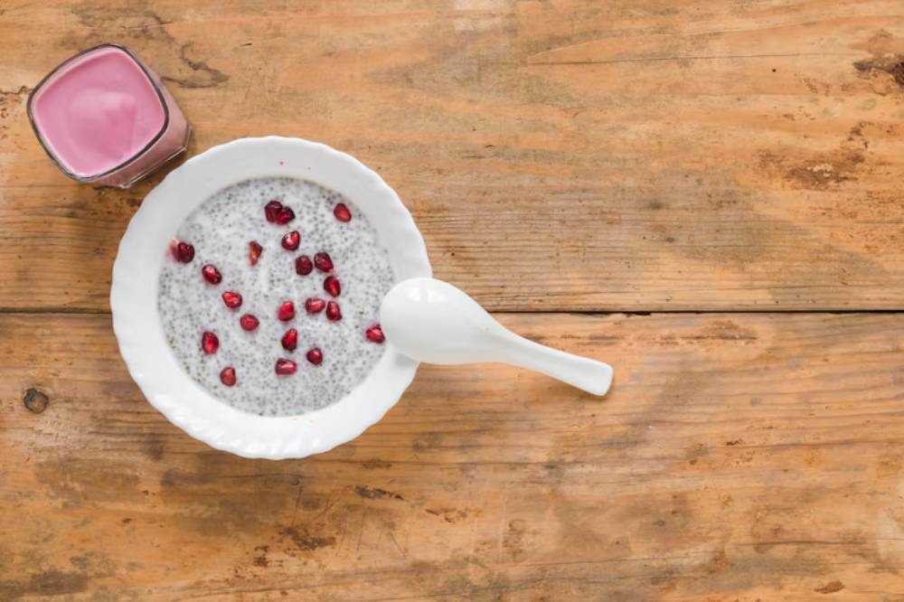 A bowl of chia pudding topped with pomegranate seeds, accompanied by a pink yogurt drink, on a wooden table.