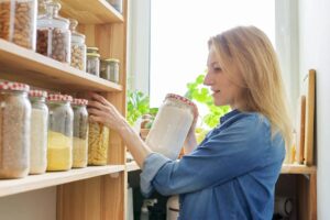 A person in a blue shirt reaches for a jar on a wooden shelf filled with assorted jars of grains, nuts, and pasta near a window.