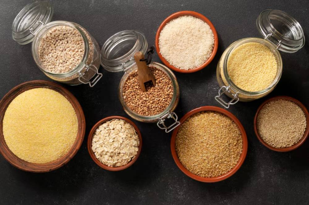 Various grains and seeds displayed in jars and bowls on a dark surface, including rice, oats, and millet.