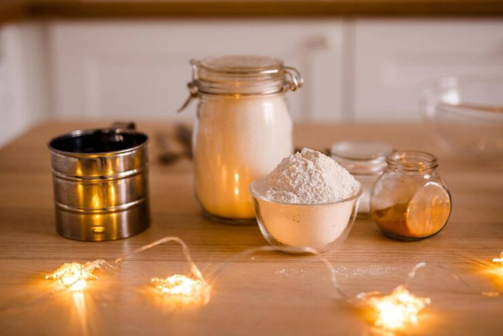 A wooden countertop displays jars of sugar, flour, and honey, alongside a measuring cup and twinkling fairy lights.
