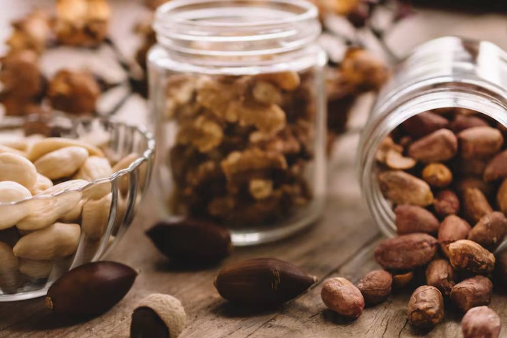Assorted nuts in glass jars and a bowl on a wooden surface, surrounded by acorns and dried flowers.