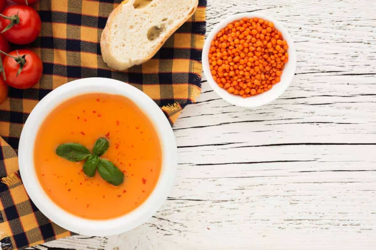 A bowl of orange soup garnished with basil, alongside cherry tomatoes, sliced bread, and a dish of orange lentils on a checkered cloth.