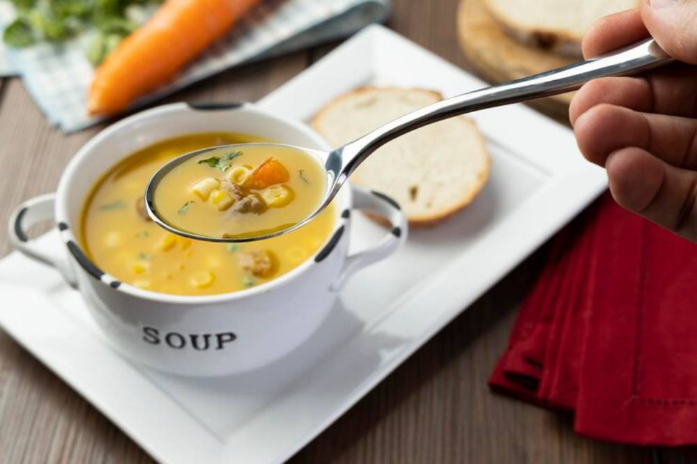 A spoonful of vibrant vegetable soup is lifted from a white bowl labeled 'SOUP,' with a slice of bread and a carrot in the background.