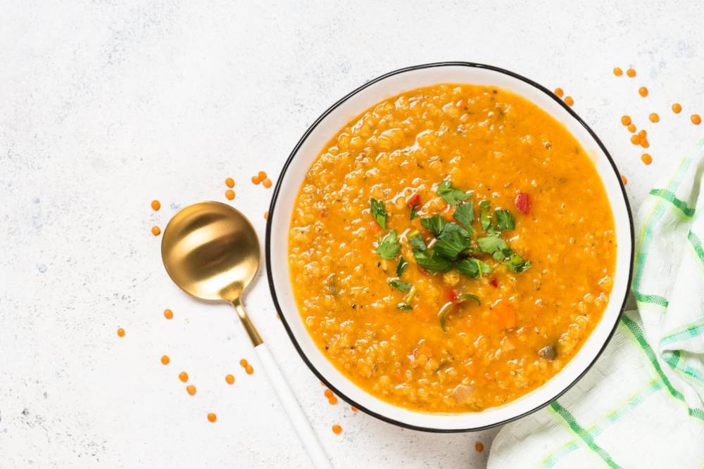 A bowl of vibrant orange lentil soup garnished with fresh herbs, accompanied by a gold spoon and scattered lentils.