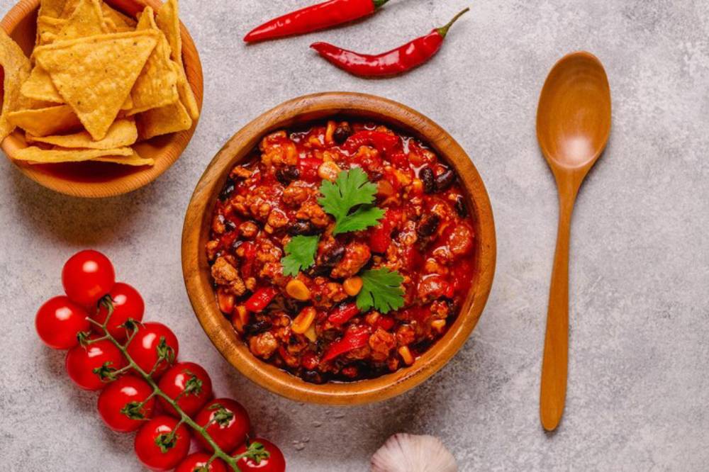 A bowl of hearty chili topped with cilantro, surrounded by tortilla chips, cherry tomatoes, garlic, and red chilies on a gray countertop.