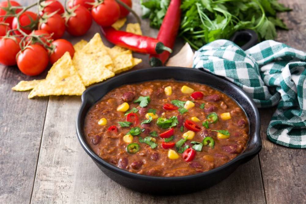 A flavorful pot of chili with beans, corn, and peppers, surrounded by fresh tomatoes, tortilla chips, and herbs on a rustic wooden table.