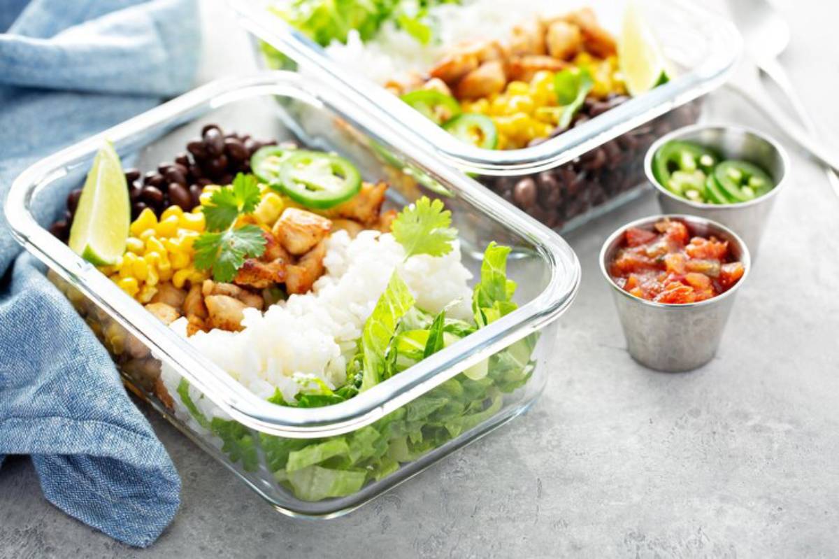 Two glass meal prep containers filled with rice, grilled chicken, black beans, corn, and fresh veggies, alongside salsa and jalapeños.