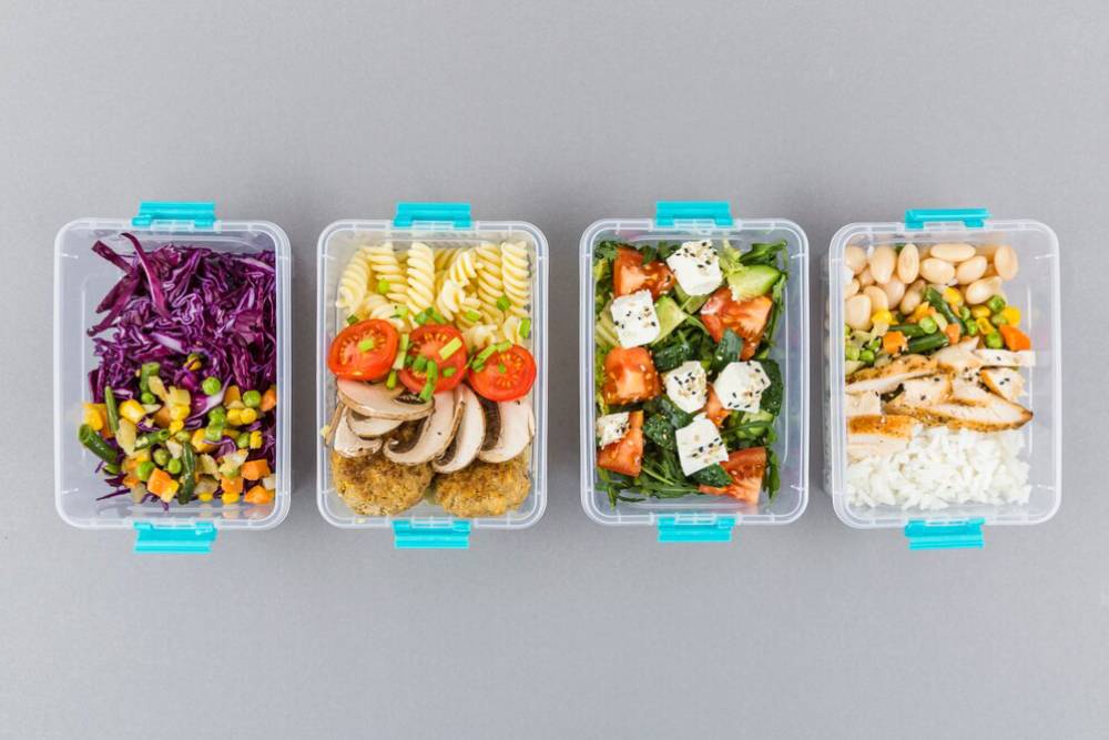 Four meal prep containers with colorful salads and proteins: purple cabbage, pasta with veggies, leafy greens with feta, and rice with chicken.