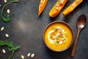 Bowl of vibrant pumpkin soup garnished with cream, herbs, and seeds, next to pumpkin wedges and a wooden spoon on a dark background.