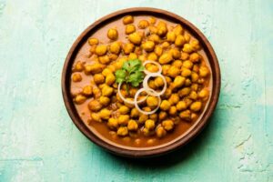 A wooden bowl filled with spicy chickpeas garnished with fresh coriander and onion rings, set against a turquoise background.