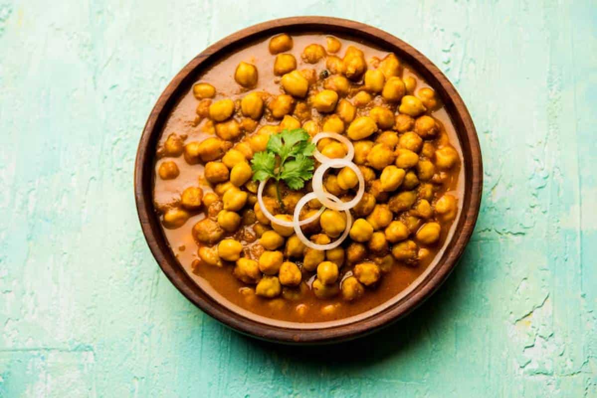 A wooden bowl filled with spicy chickpeas garnished with fresh coriander and onion rings, set against a turquoise background.