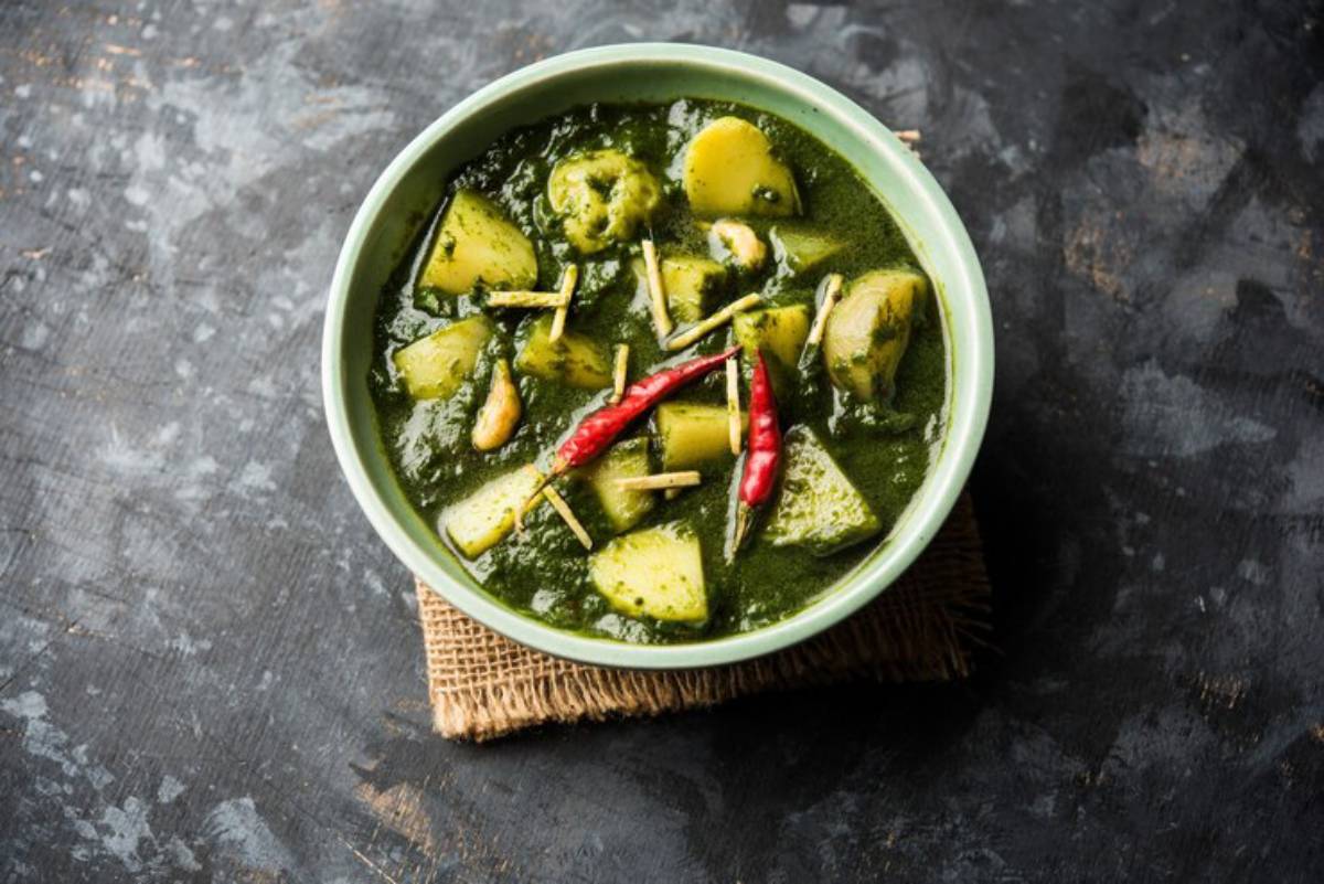 A bowl of vibrant green curry with potato chunks, spices, and garnished with red chilies and ginger, served on a burlap mat.