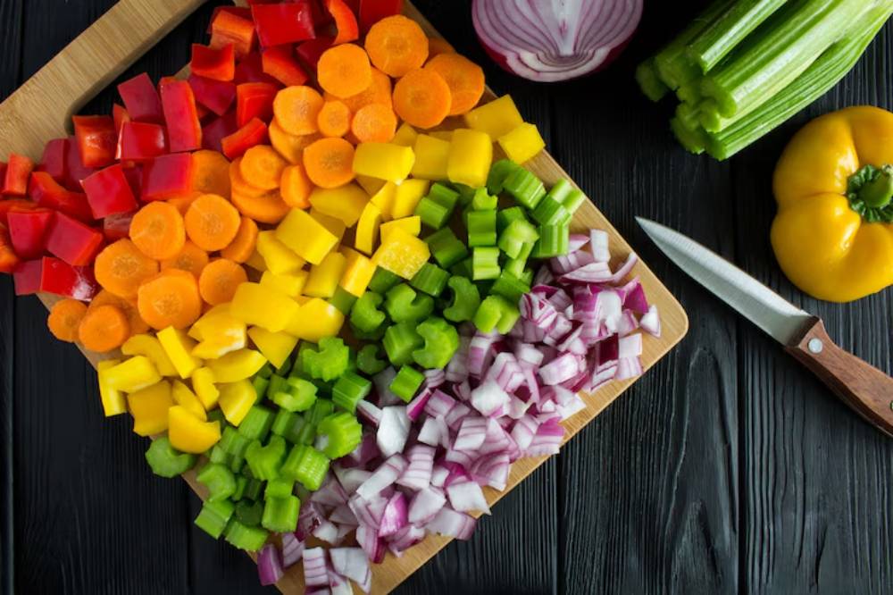 A vibrant assortment of chopped vegetables including red bell peppers, carrots, yellow bell peppers, celery, and red onions on a wooden cutting board.