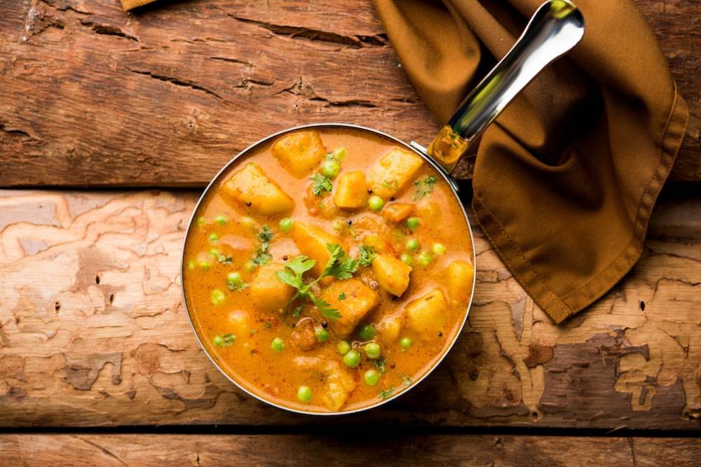 A bowl of creamy vegetable curry with chunks of potatoes and green peas, garnished with fresh cilantro, on a rustic wooden table.