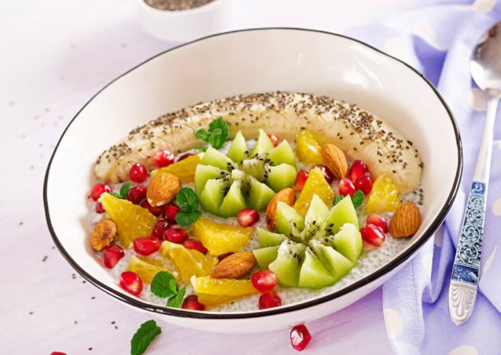 A bowl of chia pudding topped with a sliced banana, kiwi flowers, orange segments, pomegranate seeds, whole almonds, and fresh mint leaves, presented on a white plate with a black rim and a decorative spoon beside it.
