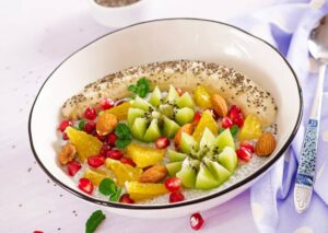 A bowl of chia pudding topped with a sliced banana, kiwi flowers, orange segments, pomegranate seeds, whole almonds, and fresh mint leaves, presented on a white plate with a black rim and a decorative spoon beside it.