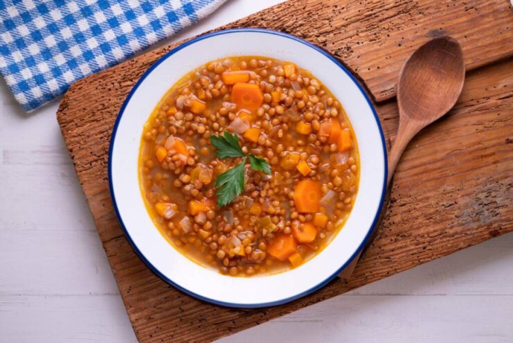 Bowl of lentil soup with carrots and herbs, served on a wooden board, alongside a blue and white checkered cloth.