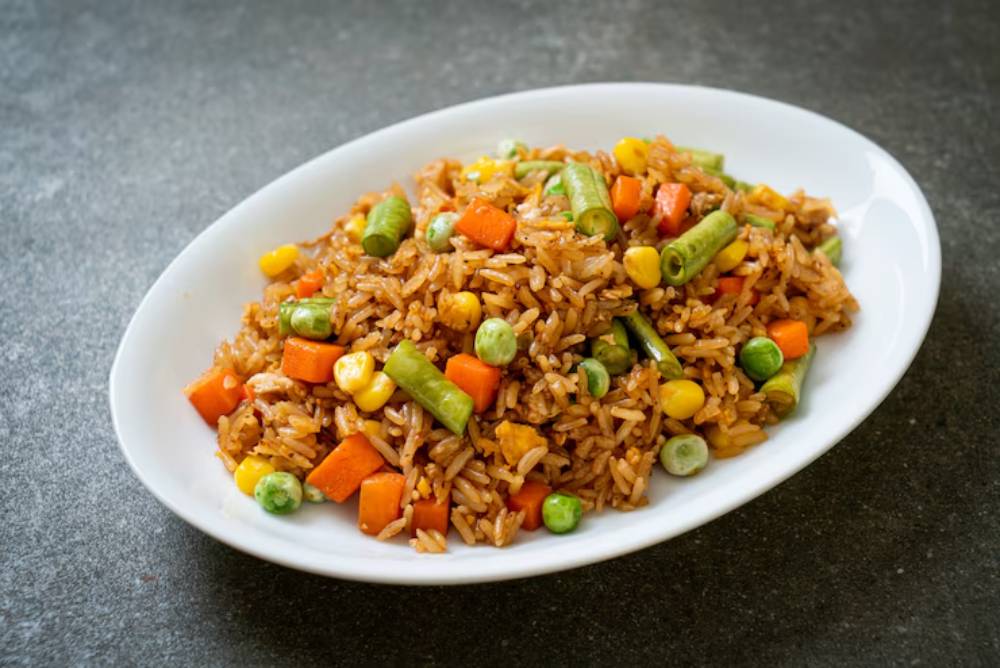 A white oval plate filled with colorful fried rice, featuring green beans, corn, carrots, and peas, set against a dark, textured background.