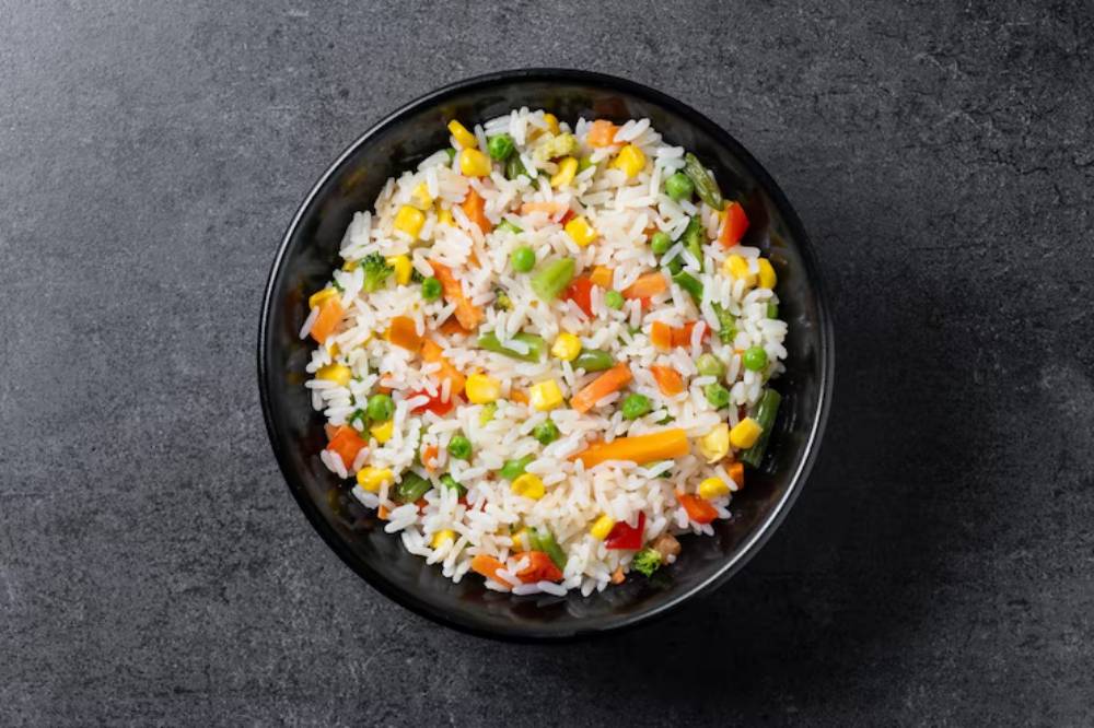 A black bowl filled with colorful vegetable fried rice, featuring peas, corn, red and yellow bell peppers, and broccoli on a dark surface.
