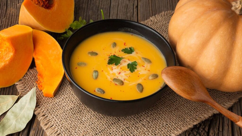 A black bowl of creamy pumpkin soup topped with pumpkin seeds and cilantro, surrounded by fresh pumpkin halves and bay leaves.
