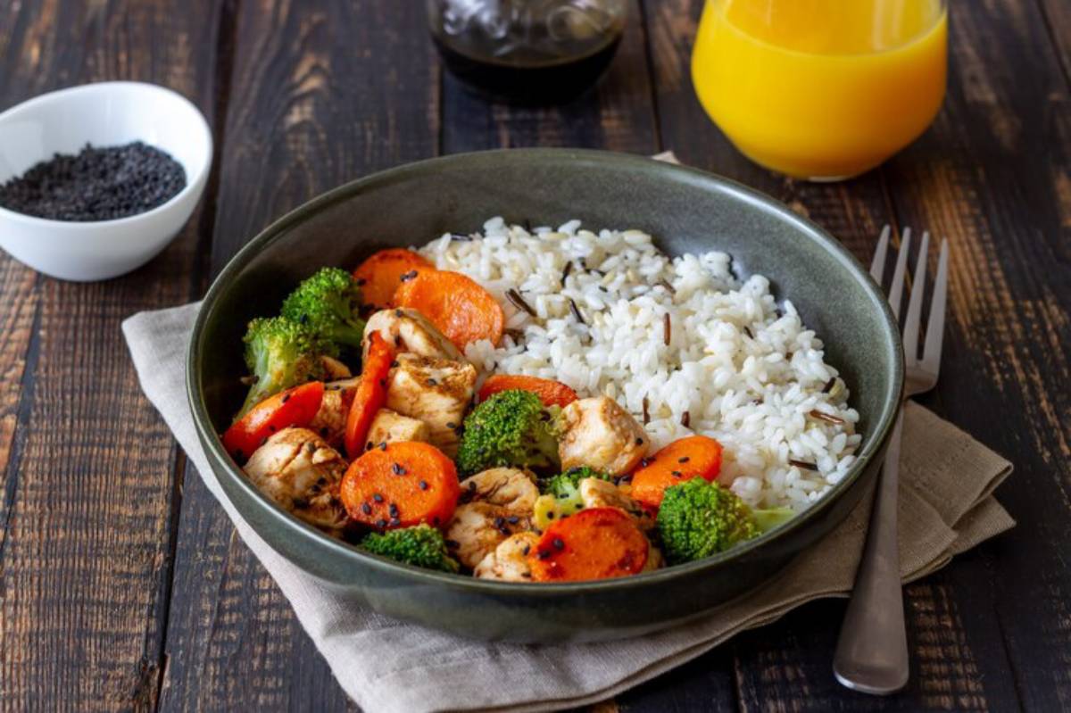 A bowl of  broccoli, and carrots served with fluffy white rice, accompanied by a side of black sesame seeds and orange juice.