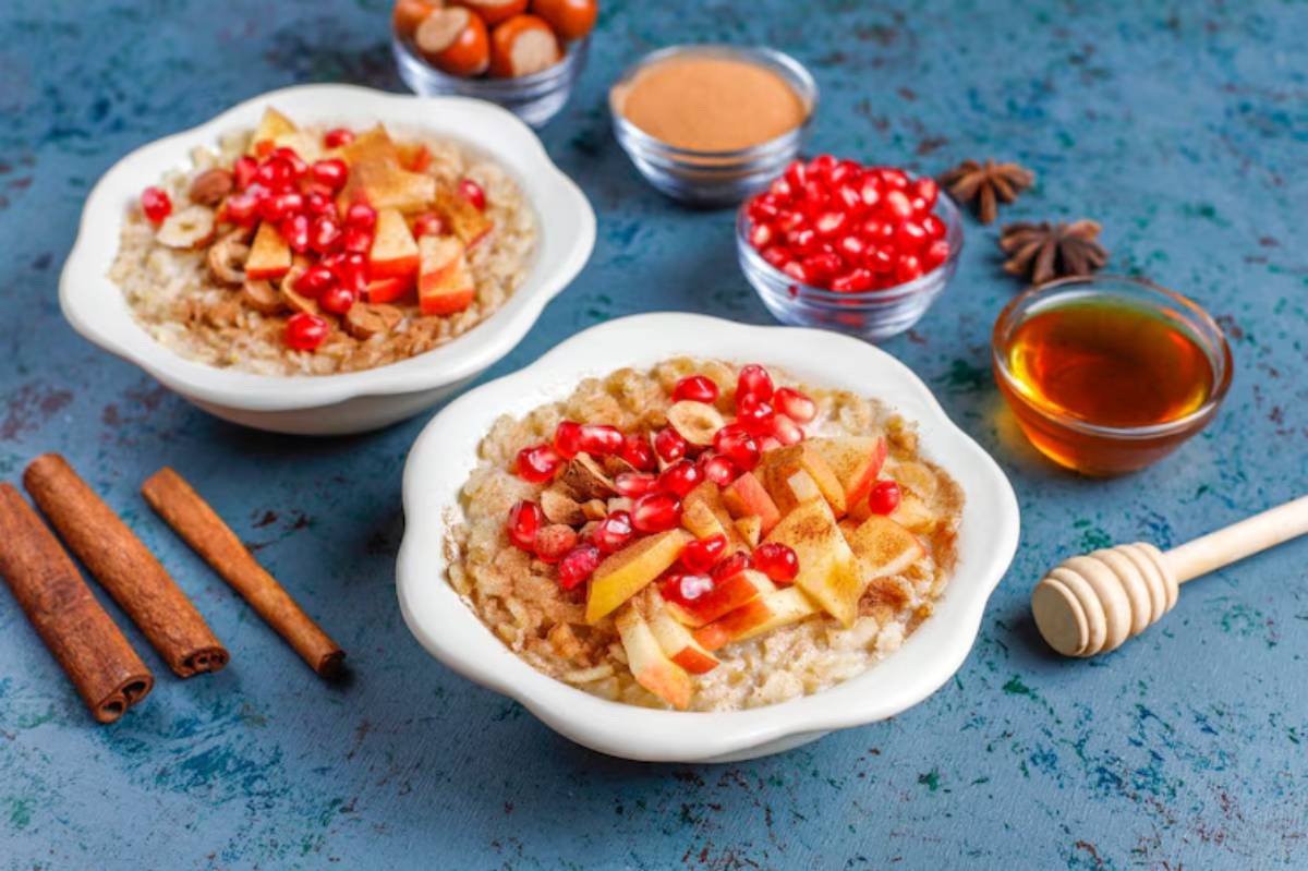 Two bowls of oatmeal topped with apples, pomegranate seeds, nuts, and cinnamon, accompanied by honey and cinnamon sticks.