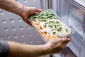 A person placing a tray of frozen meal, featuring fish, mixed vegetables, and herbs, into a refrigerator.
