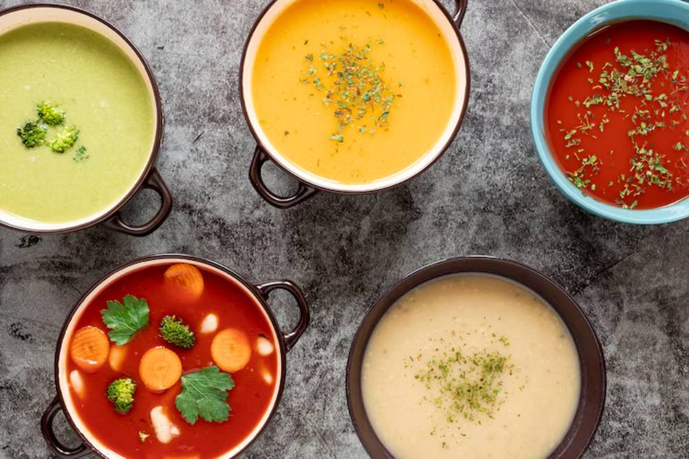 A variety of colorful soups in bowls: green broccoli, yellow cream, tomato with veggies, white chowder, and a rich red broth on a textured surface.