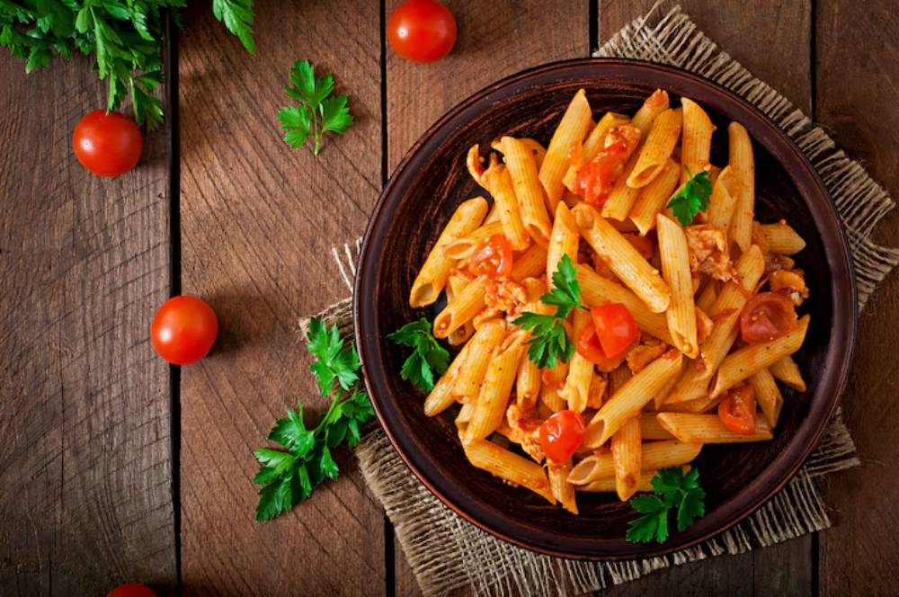 A bowl of penne pasta with cherry tomatoes and fresh parsley on a rustic wooden table, garnished with herbs.