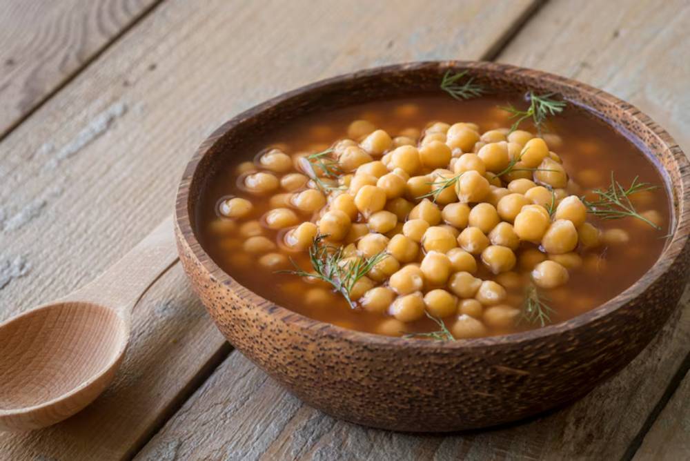 A wooden bowl filled with chickpea soup, garnished with fresh herbs, placed on a rustic wooden surface beside a wooden spoon.