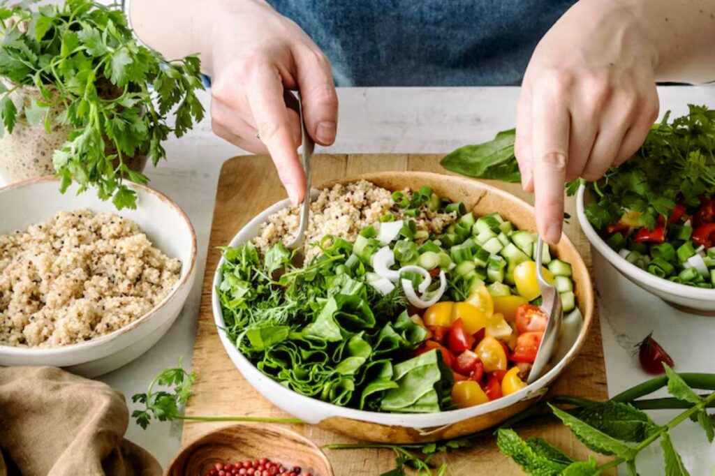 A person mixing a colorful salad with leafy greens, tomatoes, cucumbers, and quinoa on a wooden table, surrounded by fresh herbs.