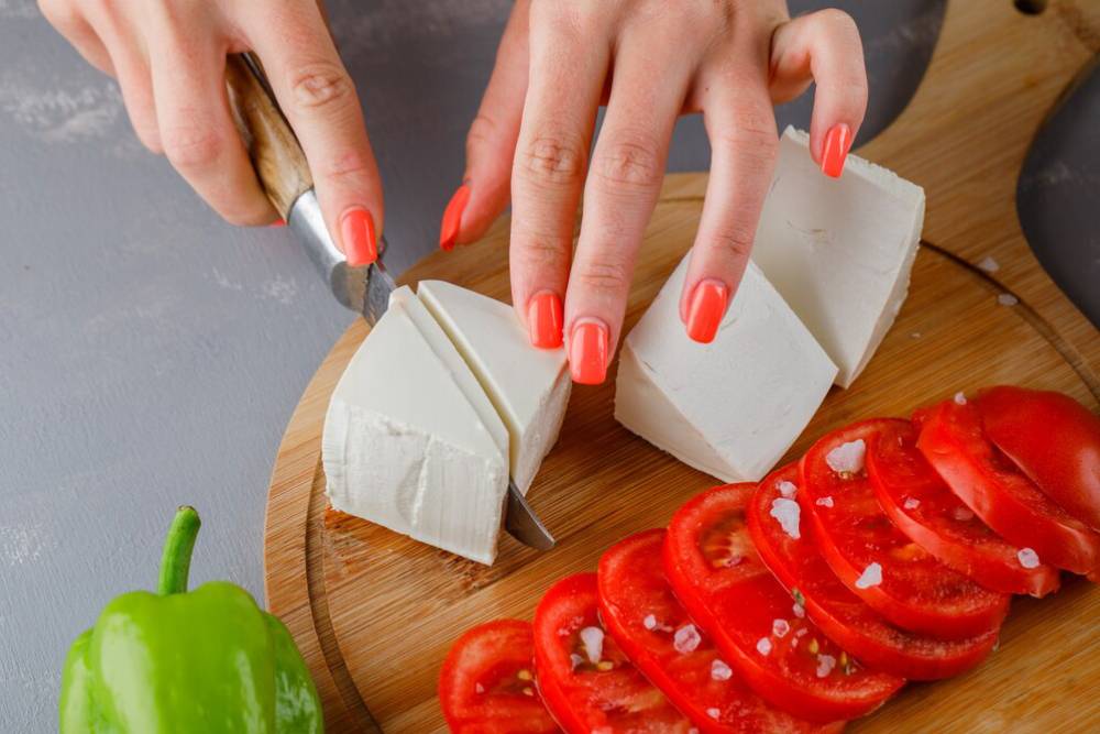 A person's hand with coral nails slices a block of white cheese on a wooden cutting board beside freshly sliced tomatoes and green pepper.