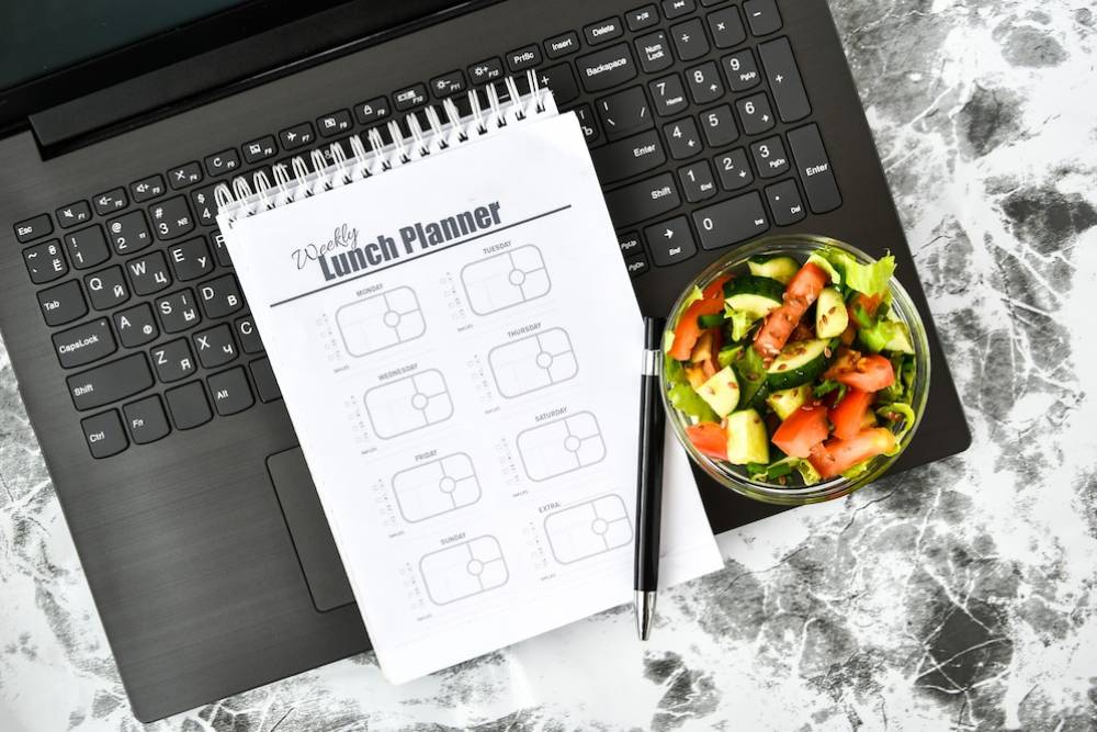 A laptop with a weekly lunch planner and a bowl of fresh salad sits atop a marbled surface, accompanied by a pen.