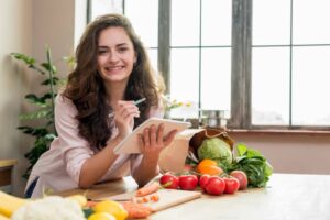A person sits at a table filled with fresh vegetables and fruits, holding a tablet and a pen, looking thoughtfully at their notes.