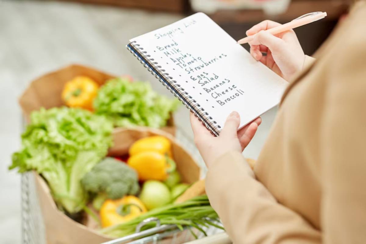 A person holds a shopping list with items like bread, milk, and vegetables, next to a cart filled with fresh produce.