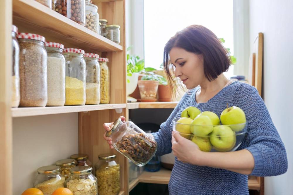 A woman in a cozy kitchen holds a bowl of green apples while inspecting a jar of nuts, with shelves of neatly stored ingredients behind her.