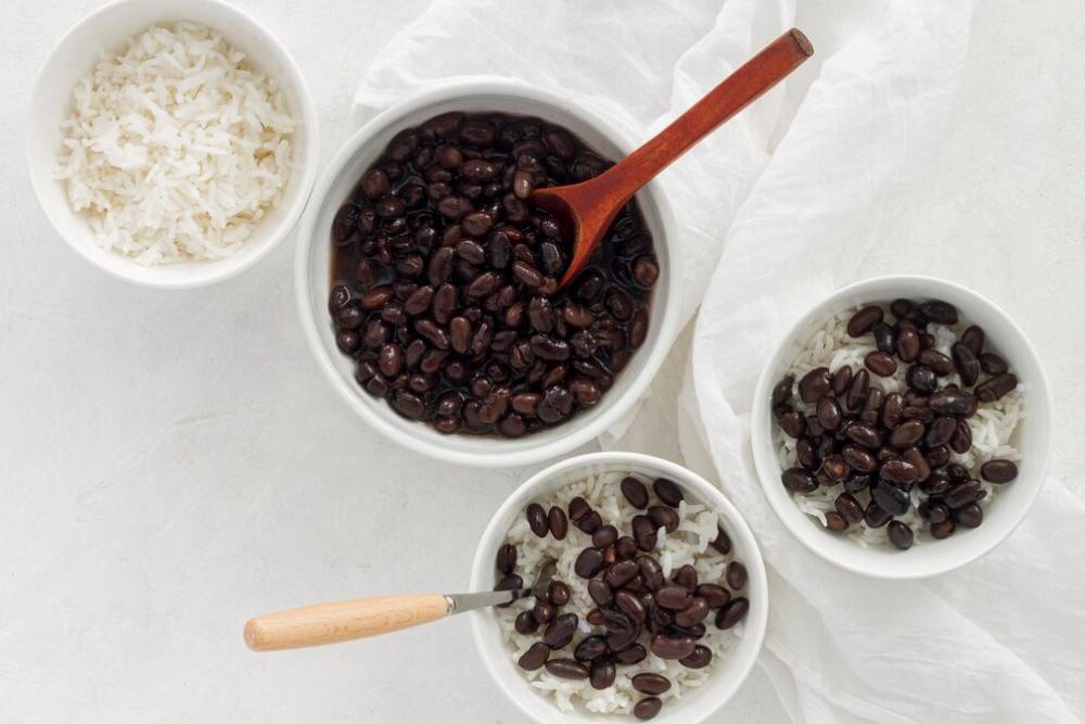 Three white bowls with rice and black beans, one with a wooden spoon, on a light background with a soft cloth.