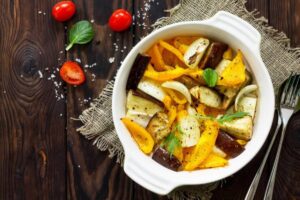 A white bowl filled with roasted vegetables, including yellow peppers, eggplant, and onions, garnished with herbs, on a wooden table.