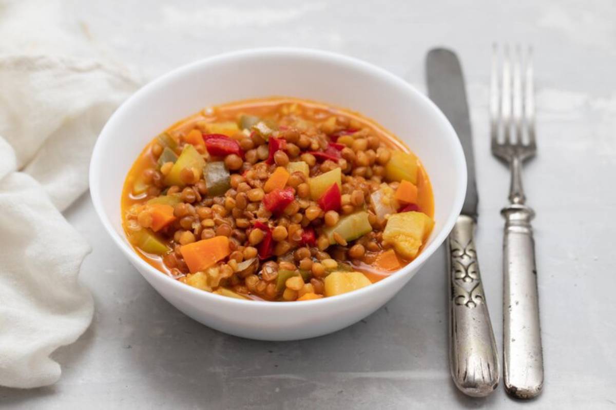A bowl of colorful lentil soup with assorted vegetables, accompanied by silver cutlery and a soft white cloth.