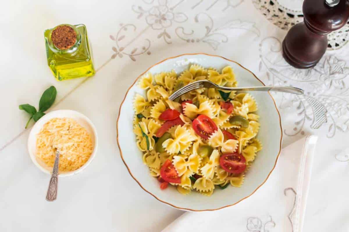A bowl of bowtie pasta with cherry tomatoes and green olives, garnished with basil, alongside olive oil and grated cheese.