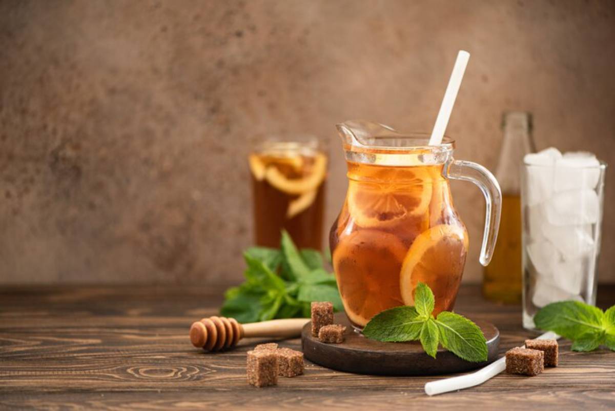 A glass pitcher of iced tea with lemon slices, mint leaves, honey, and brown sugar cubes, set against a rustic background.