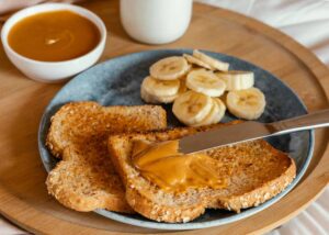 A plate with two slices of toast spread with peanut butter, banana slices, and a bowl of peanut butter on a wooden tray.