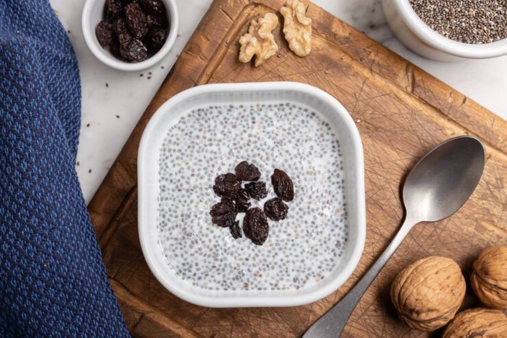 A bowl of chia pudding topped with raisins, surrounded by walnuts, a silver spoon, and a blue textured towel on a wooden cutting board.
