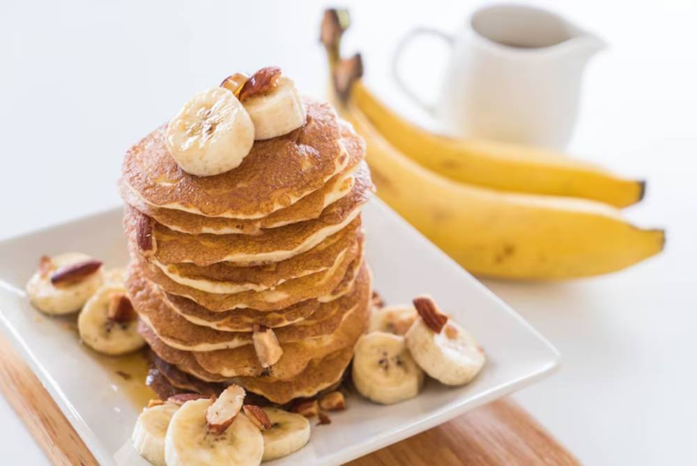 Stack of banana pancakes topped with banana slices and nuts, surrounded by whole bananas and a small pitcher in the background.