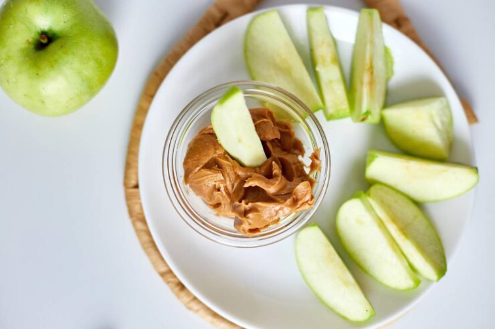 Sliced green apples on a plate with a jar of nut butter  in the center, topped with an apple slice, and a whole apple beside it.